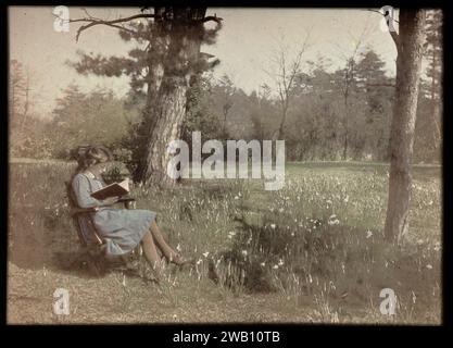 Porträt einer jungen Frau auf einem Stuhl, möglicherweise Elise Burdet, 1907 - 1930 Foto. Farbfolie Junge Frau (in einem blauen Kleid gekleidet) sitzt auf einem Stuhl, liest in einem Buch (?), auf einem Feld mit Narzissen zwischen Bäumen Niederlande Glasrutsche Stockfoto