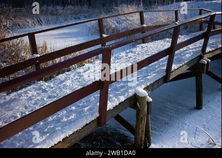 Schneebedecktes Holzfahrrad und Wanderbrücke mit Geländer über einen gefrorenen Graben in einem Wald Stockfoto