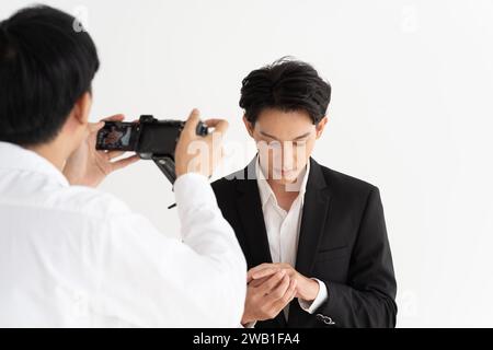 Asiatischer Bräutigam in einem stilvollen Anzug passt seine Manschettenknöpfe sorgfältig an, während ein Fotograf den Moment festhält. Eleganz im Fokus: Ein professioneller Fotograf fotografiert das Porträt eines Bräutigams Stockfoto