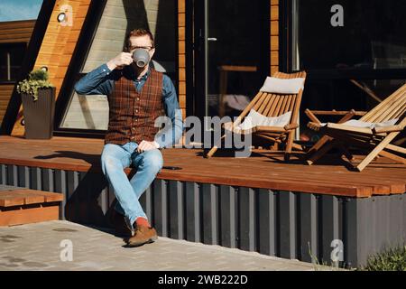 Mann mittleren Alters, der auf der Terrasse einer Holzhütte sitzt und morgens Tee trinkt Stockfoto