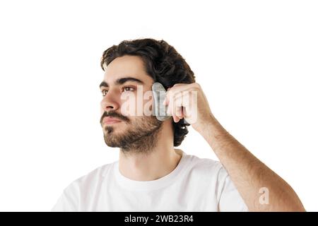 Nahaufnahme Porträt des Mannes bewegt sich sanft über die Gesichtshaut mit Massagewerkzeug vor weißem Studiohintergrund. Konzept der Gesichtspflegeroutine. Stockfoto