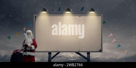 Der Weihnachtsmann überprüft eine Liste und liefert Geschenke. Er steht neben einem Plakatschild mit leeren Plakaten Stockfoto