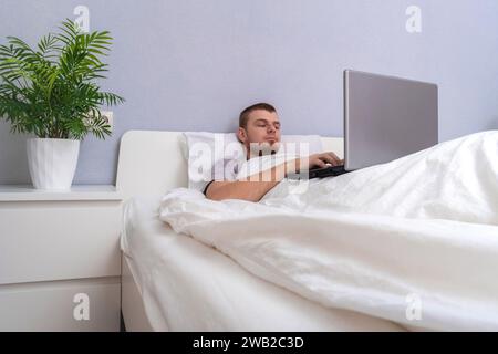 Ein Mann mit Laptop liegt im Bett. Freiberufler arbeiten von zu Hause aus. Fernunterricht. Online-Shopping ohne das Haus zu verlassen. Surfen im Internet. Spielsucht. Black-Friday-Angebot. Cyber Monday. Stockfoto