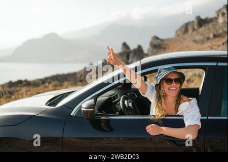 Glückliche junge Frau, die sich an das Autofenster lehnt und das Friedenszeichen mit Gesten drückt Stockfoto