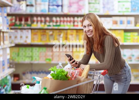 Porträt einer jungen Frau, die im Supermarkt einkaufen geht, lächelt und hält ihr Smartphone Stockfoto