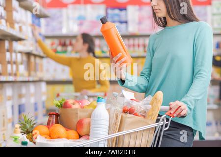 Frau, die Produkte im Lebensmittelgeschäft kauft, hält eine Waschmittelflasche und lehnt sich auf einen vollen Einkaufswagen Stockfoto