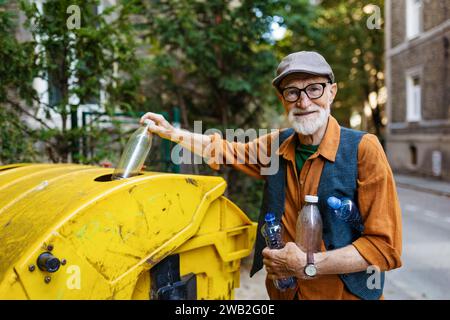 Senior Mann wirft Plastikmüll, Flaschen in den Recyclingbehälter vor der Wohnung. Ein älterer Mann sortiert den Abfall nach Material Stockfoto