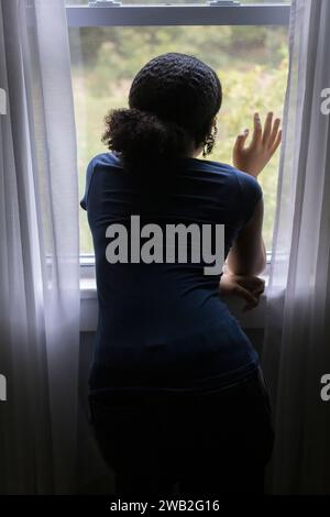 Von hinten blickt ein birassisches Teenager-Mädchen aus dem Fenster Stockfoto