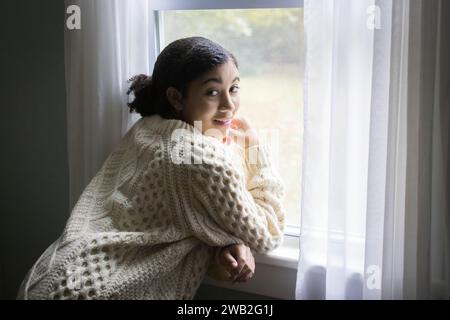 Lächelndes birassisches Teenager-Mädchen am Fenster und in die Kamera Stockfoto