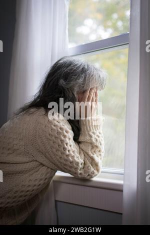 Ältere weiße Frau am Fenster mit Händen über dem Gesicht, die deprimiert aussehen Stockfoto