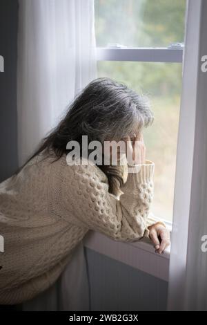 Ältere weiße Frau am Fenster mit Händen über dem Gesicht, die deprimiert aussehen Stockfoto