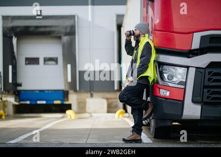 Lkw-Fahrer lehnt sich an den roten Lkw und trinkt Kaffee, wartet auf Lagerarbeiter. Stockfoto
