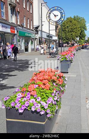 Pflanzkästen farbenfrohe Sommerblumen in langer Reihe, um Parkmöglichkeiten auf dem Bürgersteig mit Stadtuhr in der Brentwood Shoppers High Street Essex England, Großbritannien, zu blockieren Stockfoto