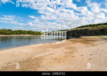 Ebbe auf einem Fluss, der eine Sandbank im Vordergrund mit einem hohen felsigen Ufer und Wald unter einem blauen, bewölkten Himmel enthüllt Stockfoto