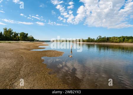 Ein Fischer steht mit einer Angelrute bei Ebbe im Wasser auf einem breiten Fluss mit bewaldeten Ufern unter blauem, bewölktem Himmel Stockfoto