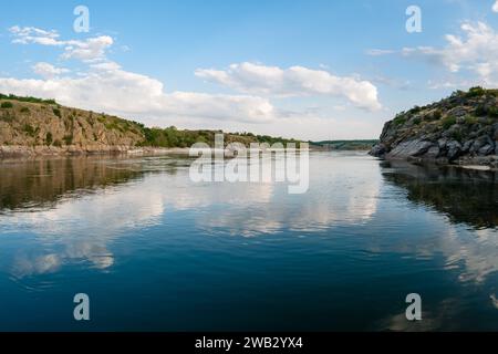 Große Kumuluswolken spiegeln sich im Wasser eines breiten Flusses mit hohen felsigen Ufern im Wald und einem langen Strand im Hintergrund wider Stockfoto