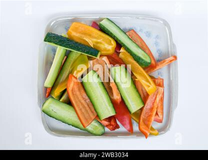 Blick von oben auf Gemüsescheiben aus gelben und roten Paprika und Gurken auf einer quadratischen Porzellanschale auf weißem Hintergrund Stockfoto