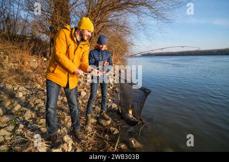 Vater und Sohn fischen an sonnigen Wintertagen. Sie haben einen Fisch gefangen und halten ihn in einem Landenetz. Stockfoto