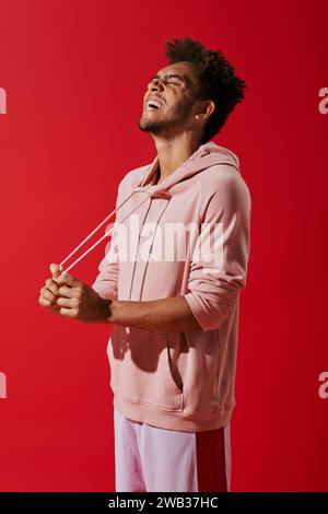 Glücklicher afroamerikanischer Mann, der Kordelzüge aus rosa Hoodie zieht und lacht auf rotem Hintergrund Stockfoto