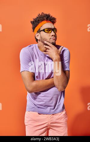 Nachdenklicher junger afroamerikanischer Mann in Stirnband und Sonnenbrille auf orangem Hintergrund, lustiges Gesicht Stockfoto