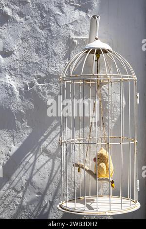 Raue Wand eines Hauses mit einer weißen Fassade in hellem Sonnenlicht. Alter Vogelkäfig mit einem gehäkelten gelben kleinen Vogel - Papagei darin. Teguise, Lanzarote, Stockfoto