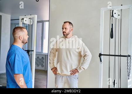 Ernsthafter bärtiger Mann mit Hüfthänden, der jungen Spezialisten im Kinesiotherapiezentrum hört Stockfoto