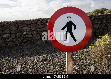 Verkehrsschild ohne Fußgänger. Aufkleber mit Gesicht und drei Augen. Kies im Hintergrund und ein Steinzaun. Bewölkter Himmel. Lanzarote, Can Stockfoto