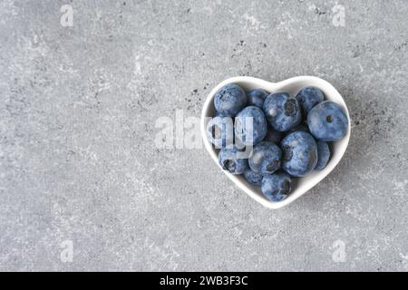 Frische Heidelbeeren in einer kleinen herzförmigen Schüssel auf grauem Betonhintergrund mit Kopierraum. Bio-Beeren, gesundes Essen, wilde Beeren. Draufsicht, fla Stockfoto