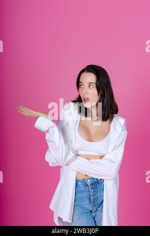 Schockierte junge Dame in weißem Hemd und Bralette mit einer ablehnenden Handgeste, rosa Hintergrund. Stockfoto