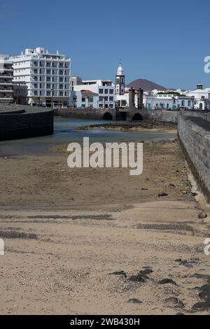 Steinbrücke vom Schloss von St. Gabriel in die Stadt. Weiße moderne Gebäude und ein Kirchturm an der Küste des Atlantischen Ozeans. Arrecife, La Stockfoto