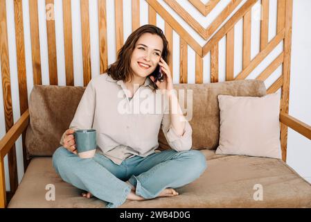 Fröhliche junge Frau, die am Telefon spricht, sich auf dem Sofa entspannt, während der Freizeit erfrischende Getränke genießt. Fröhliche junge Frau ruft telefonisch an, spricht Stockfoto