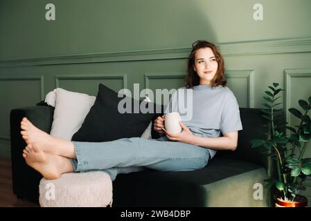 Eine nachdenkliche junge Frau mittleren Alters, die sich ausruhen und eine Tasse Tee auf der Couch genießt, nachdenkt und auf den offenen Raum blickt. Nachdenkmoment. Aufmerksam Stockfoto