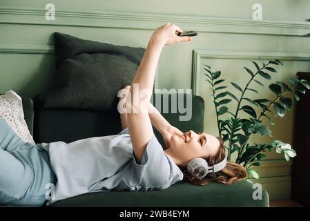 Glückliche junge Frau, die schnurlose Kopfhörer trägt, Musik auf der Couch hört, Spaß hat und Selfies macht. Musikalische Freude. Die Junge Frau Fängt Spaß Ein Stockfoto