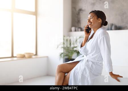 Fröhliche schwarze Frau, die Bademantel trägt und sich im Badezimmer auf dem Handy unterhalten kann Stockfoto