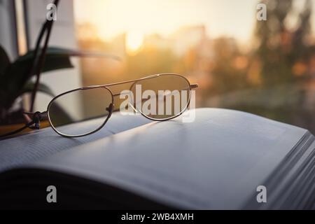 Lesebrille und ein offenes Buch auf der Fensterbank vor dem Hintergrund der Sonne. Home Holiday Konzept Stockfoto