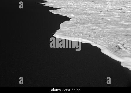 Meist verschwommener schwarzer Sandstrand mit weißem Schaum aus Meereswellen Hintergrund Stockfoto