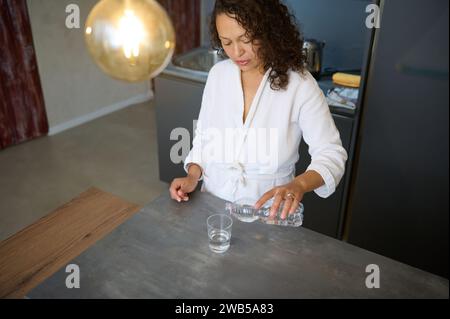 Blick von oben auf eine selbstbewusste junge Frau in weißem Bademantel, die an der Küchenzeile in einem modernen minimalistischen Wohnraum steht und etwas Wasser in einen gießt Stockfoto