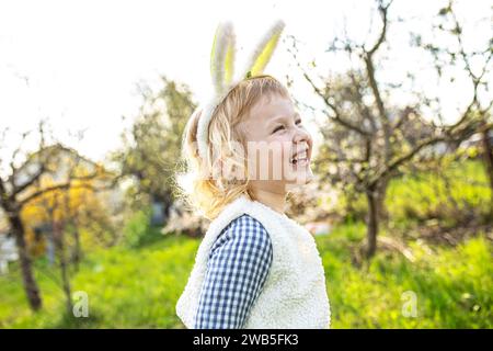 Glückliches kleines Mädchen, das Hasenohren trägt und auf grünem Gras im Garten läuft, um Ostern zu feiern Stockfoto