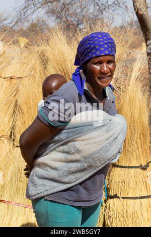 Afrikanische Frau, die das Baby im Rücken trägt, in ein Handtuch gewickelt, auf traditionelle Weise, Strohgras im Hintergrund, Dorfleben. Stockfoto