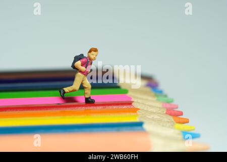 Miniatur Tiny People Spielzeug Fotografie. Ein Junge mit Schulrucksack, der über dem Buntstift läuft. Isoliert auf grauem Hintergrund. Bild Foto Stockfoto