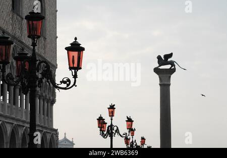 Eine wunderbare perspektivische Aussicht auf eine Reihe von Leuchtenpfosten mit roten Tönen auf dem Markusplatz von Venedig, Italien. Beachten Sie die Seegülle im Flug. Stockfoto