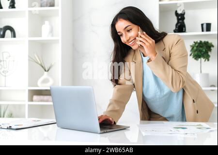 Eine geschäftige erfolgreiche, hübsche indische oder arabische Geschäftsfrau, Firmenangestellte, ceo, steht neben einem Tisch im Büro, spricht per Smartphone, führt Gespräche mit Partner oder Kunden, benutzt Laptop, lächelt Stockfoto