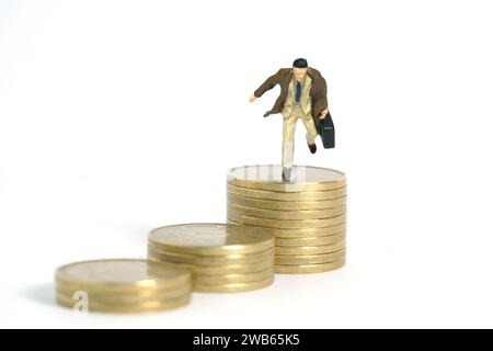 Miniatur-Kleinkinder-Spielfiguren-Fotografie. Ein Geschäftsmann, der einen Mantel trägt und über den goldenen Münzstapel springt. Isoliert auf weißem Hintergrund. Bild Foto Stockfoto