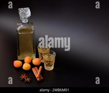 Aprikosenlikör mit Gewürzen auf schwarzem Hintergrund, starker hausgemachter Alkohol in einer Karaffe und zwei Gläsern, reifes Obst, Anis und Zimt. Nahaufnahme. Stockfoto