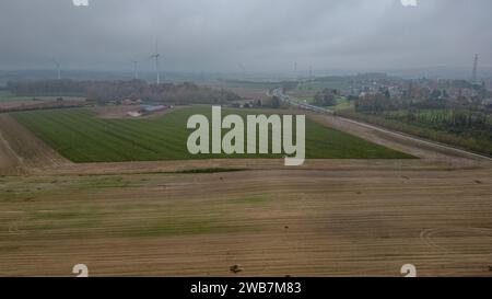 Dieses Bild zeigt eine bedeckte Tagesansicht rollender landwirtschaftlicher Felder aus der Luft. Der bewölkte Himmel wirft ein gedämpftes Licht über die Landschaft und betont die gedämpften Erdtöne der gepflügten und grünen Felder. Im Hintergrund stehen Windturbinen hoch und symbolisieren erneuerbare Energien und die Nachhaltigkeitsbemühungen der modernen Landwirtschaft. Eine Straße schlängelt sich durch die Landschaft und führt zu einer kleinen Ansammlung von Gebäuden, die auf das ländliche Leben hinweisen. Der Kontrast zwischen bearbeitetem Land und unberührten Grünflächen verdeutlicht den Flickenteppich der landwirtschaftlichen Nutzung. Die allgemeine Stimmung ist Stockfoto