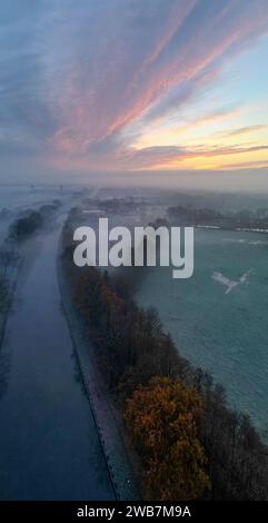 Dieses vertikale Luftbild fängt den bezaubernden Moment der Dämmerung ein, während es eine Flusslandschaft im Herzen des Herbstes beleuchtet. Ein Fluss fließt ruhig durch den Rahmen, umgeben von Bäumen, die die leuchtenden Farben des Herbstblatts zeigen. Der Himmel ist mit breiten Pinselstrichen aus rosa und orangen Wolken gemalt, die den Beginn des Tages signalisieren. Der Nebel schwebt über dem Boden und fügt eine weiche, mystische Schicht über den Feldern hinzu, und die herbstlichen Farben der Bäume bilden einen auffälligen Kontrast zu den gedämpften Tönen des umliegenden frostbedeckten Grases. Herbstliche Dämmerung am Fluss. Hohe Qualität Stockfoto