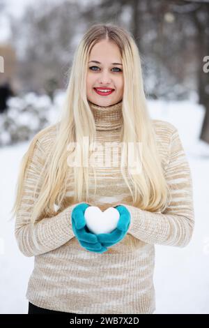 Eine junge Frau mit blondem Haar in einem Pullover hält in einem Winterpark ein Herz aus Schnee in den Händen Stockfoto