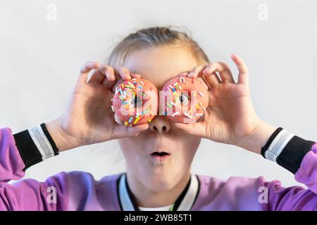 Das süße, lustige Mädchen in einem blauen T-Shirt hält knallige rosa Donuts wie eine Brille neben ihren Augen und sieht dich auf einem rosa Hintergrund an. Fröhliche Kindheit Stockfoto