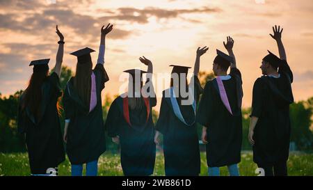 Silhouetten von Absolventen in schwarzen Gewändern, die ihre Arme gegen den Sonnenuntergang schwenken. Stockfoto