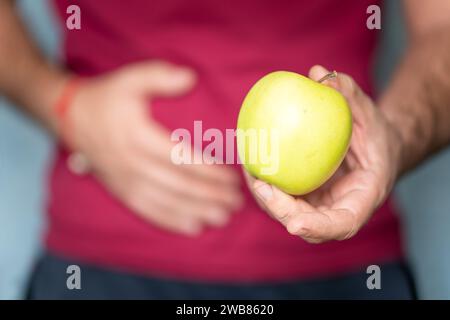 Junger Mann, der Apfel hält, der rote Hemd trägt, blauer Hintergrund mit Hand auf dem Bauch, weil Übelkeit, schmerzhafte Krankheit sich unwohl fühlt. Vergiftung durch Nein Stockfoto